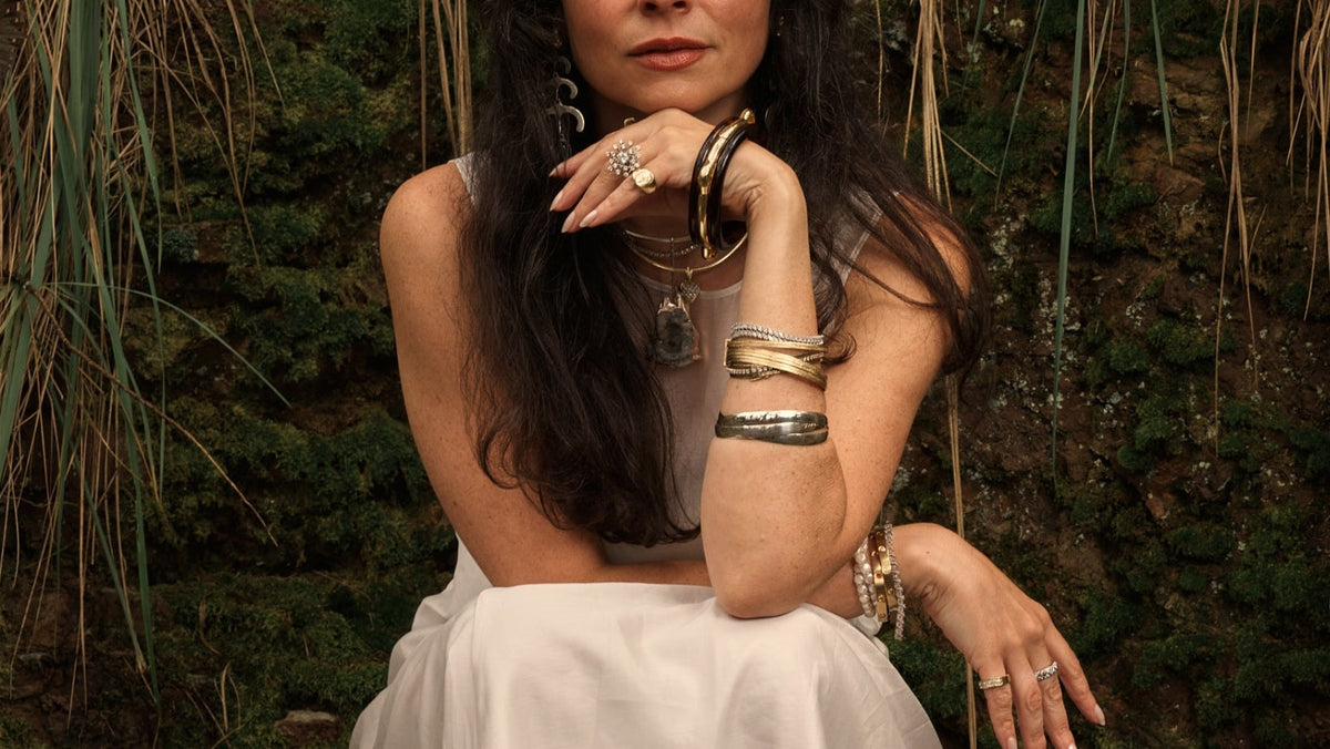 Woman in a white dress wearing stacked bangles, seated against a mossy wall